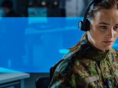 Woman with headset on, in a room with other people working on computers.